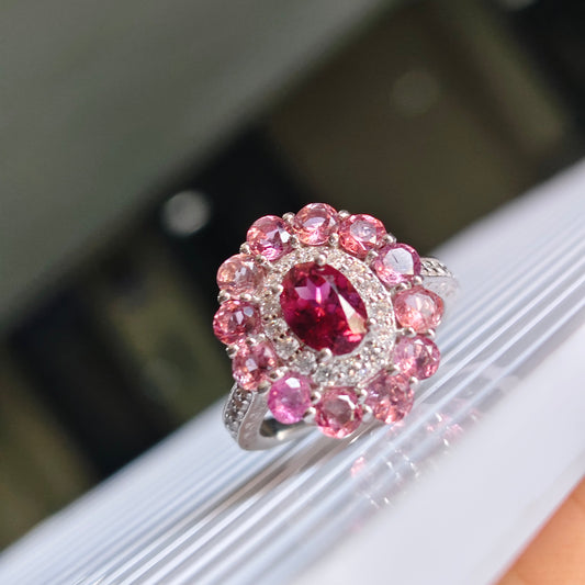 A close-up of a silver-toned ring featuring a central oval deep pink gemstone surrounded by a halo of round pink stones, displayed on a white ribbed surface.