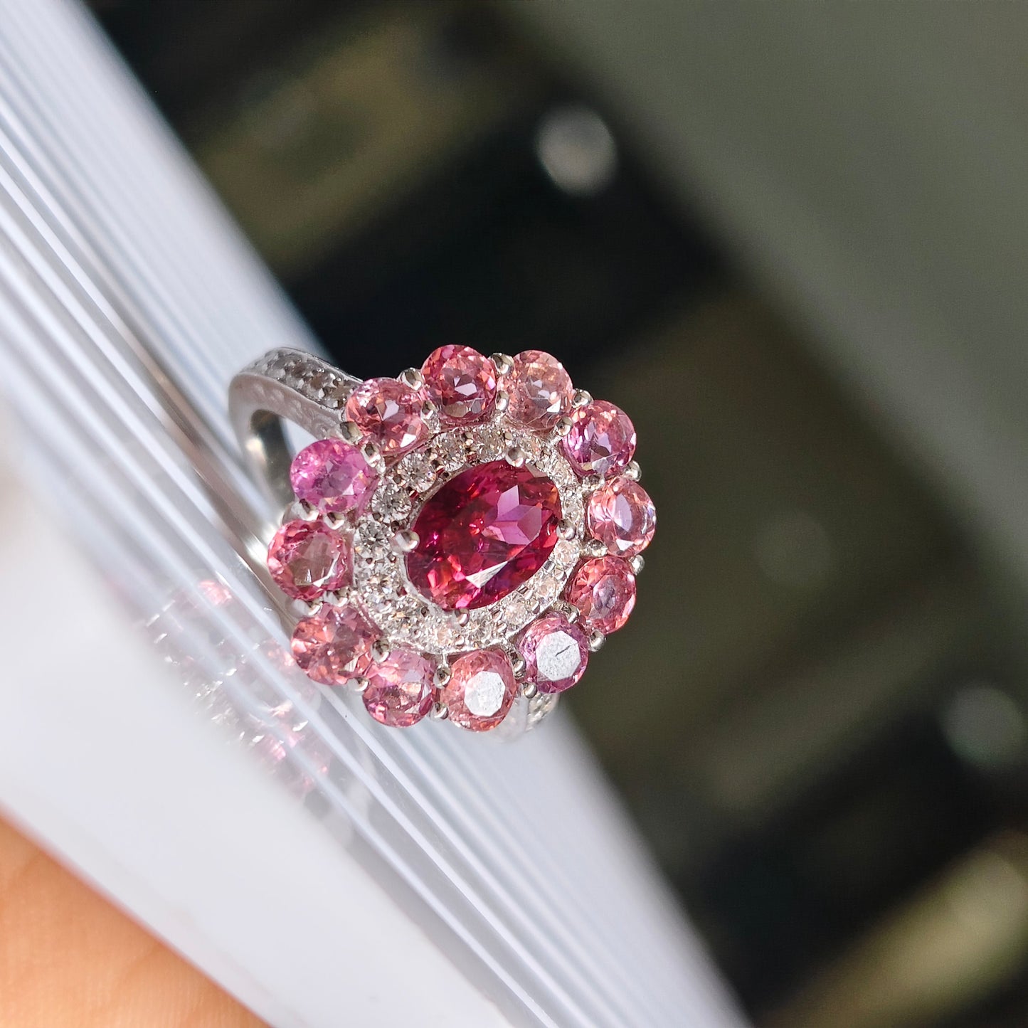 A silver ring featuring a central oval red gemstone surrounded by a halo of round pink and clear stones, resting on a white textured surface.