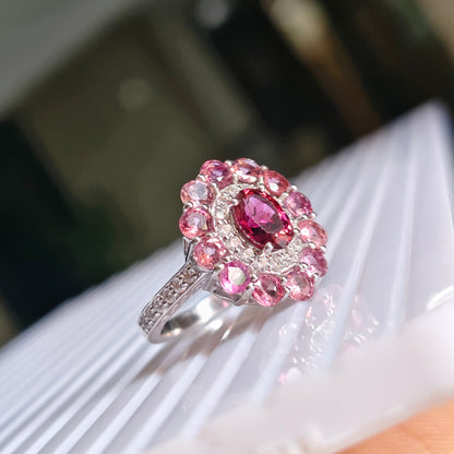 A silver-toned ring with a large oval pink gemstone surrounded by a halo of smaller pink stones, resting on a white ribbed surface.