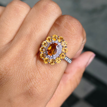 A close-up of a hand wearing a ring with a central oval amber-brown gemstone surrounded by a halo of yellow round-cut stones and small clear accent stones on a silver-toned band.
