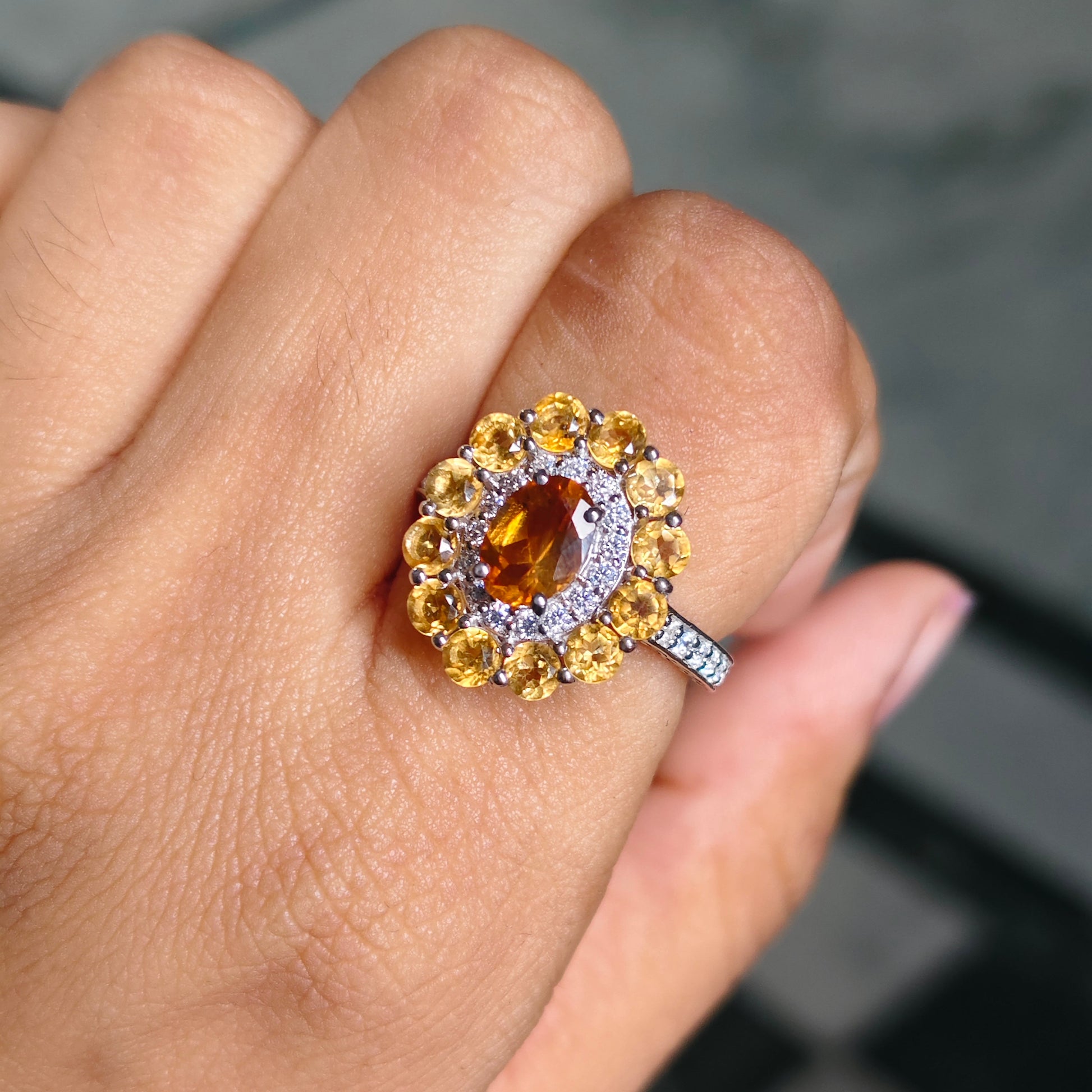 A close-up of a hand wearing a ring with a large oval amber center stone, surrounded by a halo of yellow and clear stones on a silver-toned band.