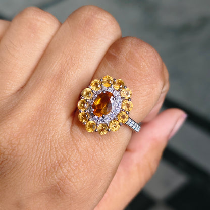A close-up of a hand wearing a ring with a large oval amber center stone, surrounded by a halo of yellow and clear stones on a silver-toned band.
