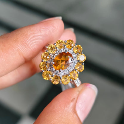 A close-up of a hand holding a silver ring with a large oval yellow center stone and multiple round yellow accent stones in a halo setting.