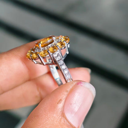 A close-up of a hand holding a silver ring with a large yellow gemstone cluster and small clear stones along the band, set against a blurred gray background.