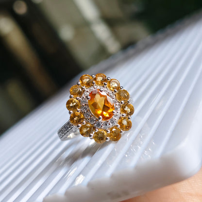 A silver ring with a large oval orange center stone and a surrounding halo of smaller round yellow-gold stones, resting on a white ribbed surface.