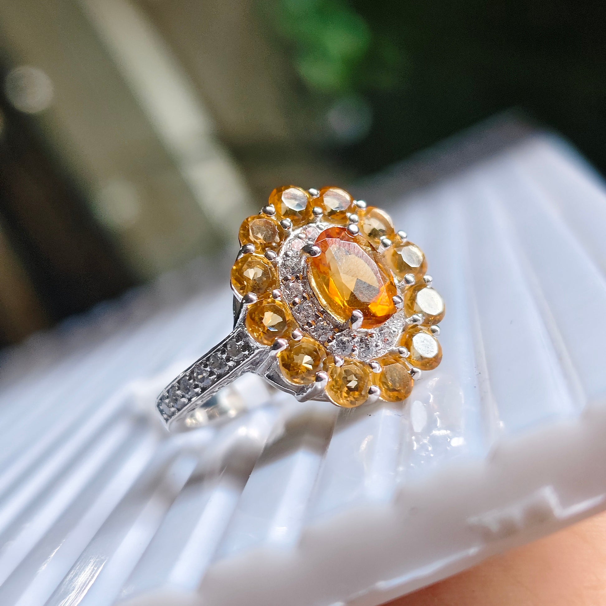 A close-up of a silver and gold ring with a central oval amber gemstone and surrounding halo accents, resting on a white textured display stand.