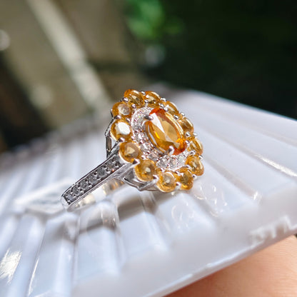 A silver ring with a large oval yellow gemstone surrounded by a halo of round yellow and clear stones, resting on a white display tray held by a hand.