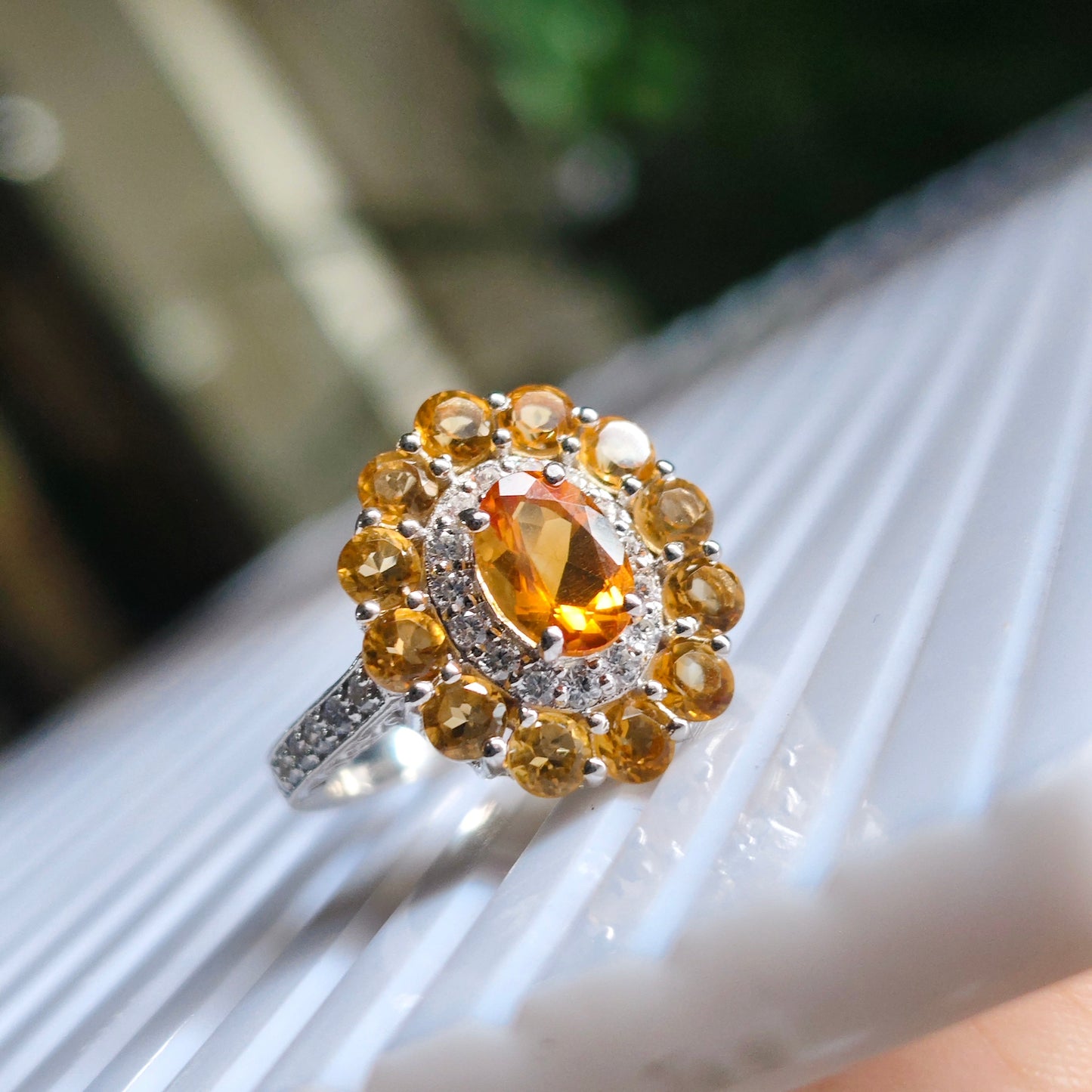 A silver ring with a large oval yellow gemstone surrounded by a halo of round yellow and clear stones, resting on a white ribbed surface.