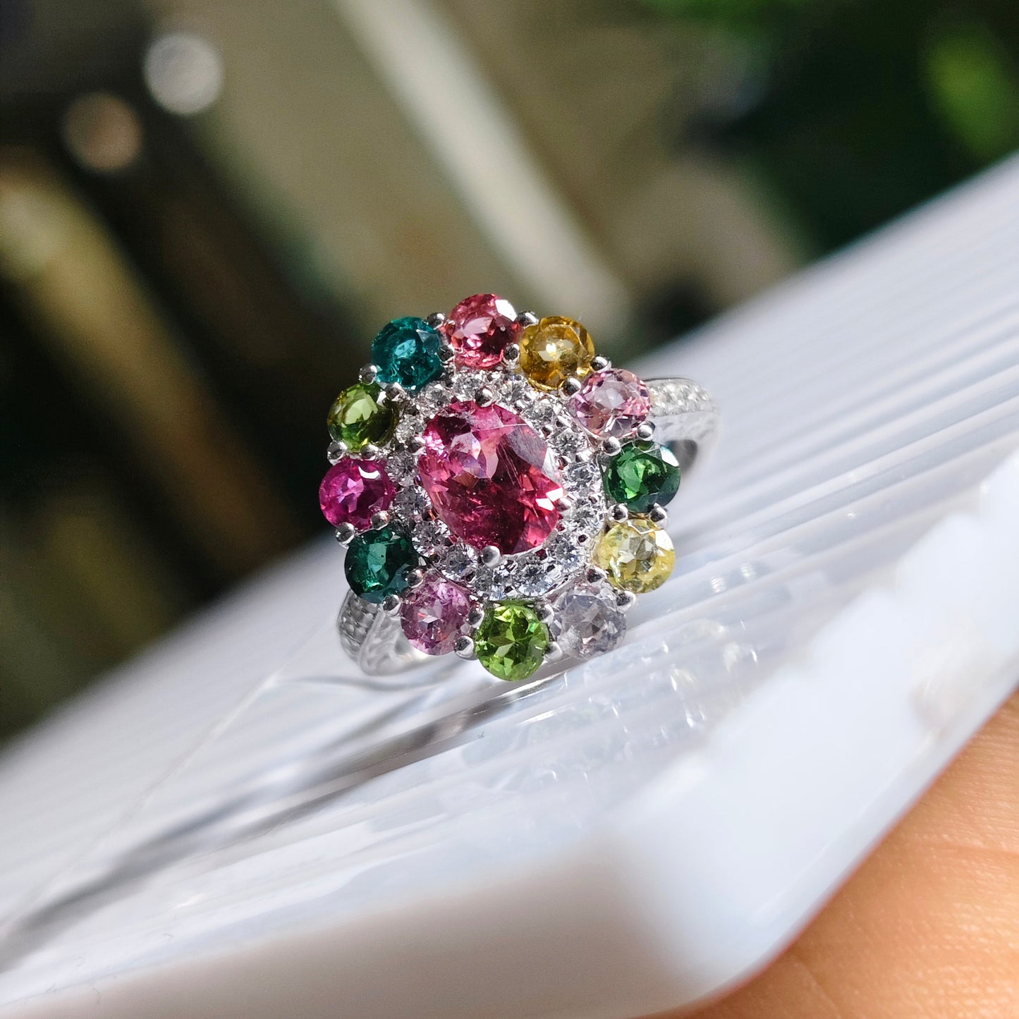 A silver-toned cluster ring with multiple round-cut multicolored gemstones, including pink, green, blue, yellow, and gold, resting on a white textured surface.