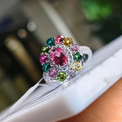 A silver-toned cluster ring with a large pink center stone and surrounding multicolored gemstones, shown resting on a white surface with a hand holding it.