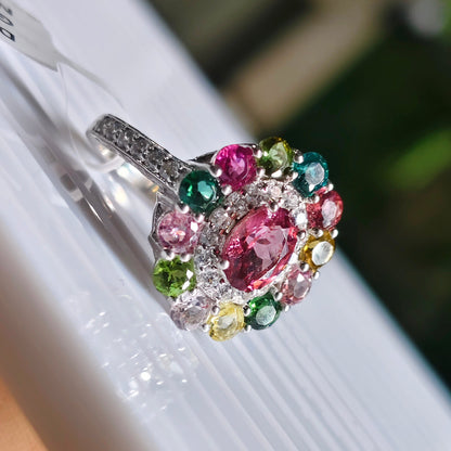 A multicolor gemstone cluster ring held by a hand on a white display card. The ring features a large oval pink center stone surrounded by smaller pink, green, yellow, and white stones in a silver-toned setting.