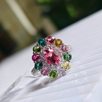 A silver-toned cluster ring featuring a central pink gemstone surrounded by multiple smaller multicolored gemstones, displayed on a white ribbed surface with a blurred green background.