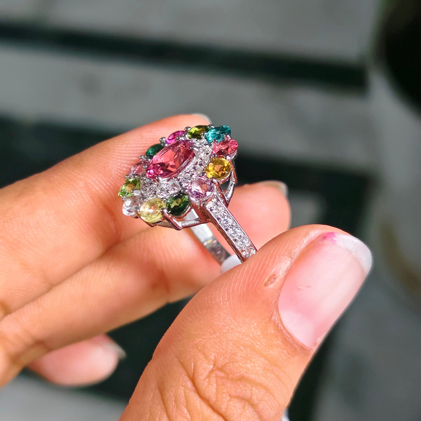 A close-up of a hand holding a colorful gemstone ring. The ring features a cluster of multicolored stones, including pink, green, yellow, and blue, set in a silver-toned metal band with small clear stones.