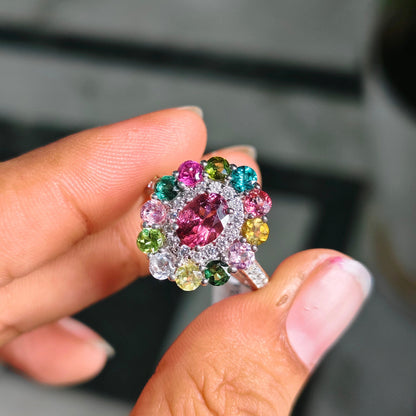 A close-up of a hand holding a colorful gemstone ring. The ring features a central pink gemstone surrounded by a cluster of multicolored stones in pink, green, blue, yellow, and orange, set in a silver-toned metal.