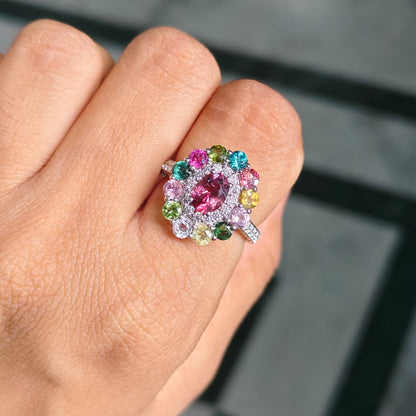 A close-up of a hand wearing a colorful flower-shaped ring with a large pink center stone surrounded by pink, blue, green, yellow, and white accent stones on a silver-toned band.