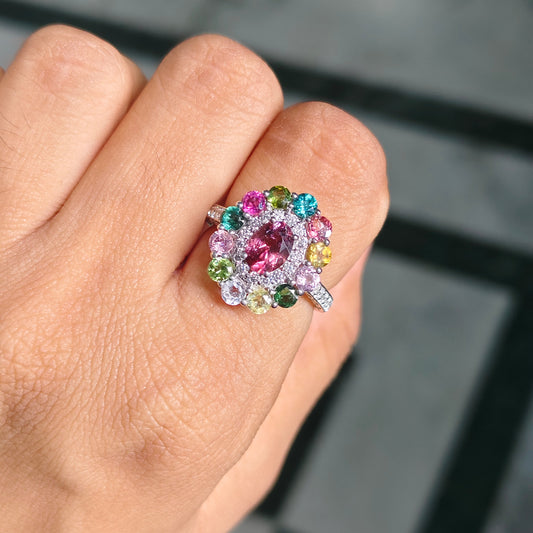 A close-up of a hand wearing a colorful flower-shaped ring with a large pink center stone surrounded by pink, blue, green, yellow, and white accent stones on a silver-toned band.