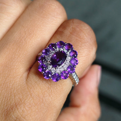 A close-up of a hand wearing a silver ring with a large oval purple amethyst center stone surrounded by a halo of smaller round purple stones and clear accent stones.