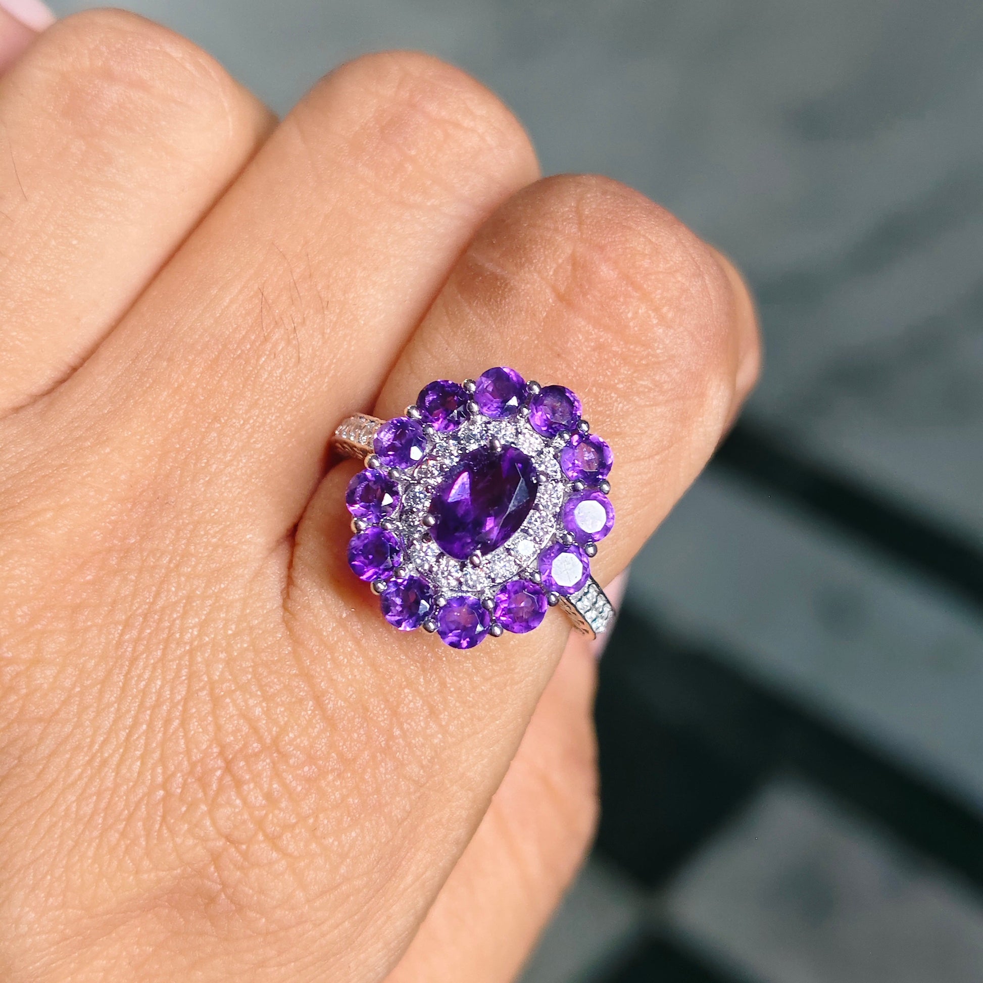 A close-up of a hand wearing a silver ring with a purple amethyst cluster setting and a central oval purple gemstone.