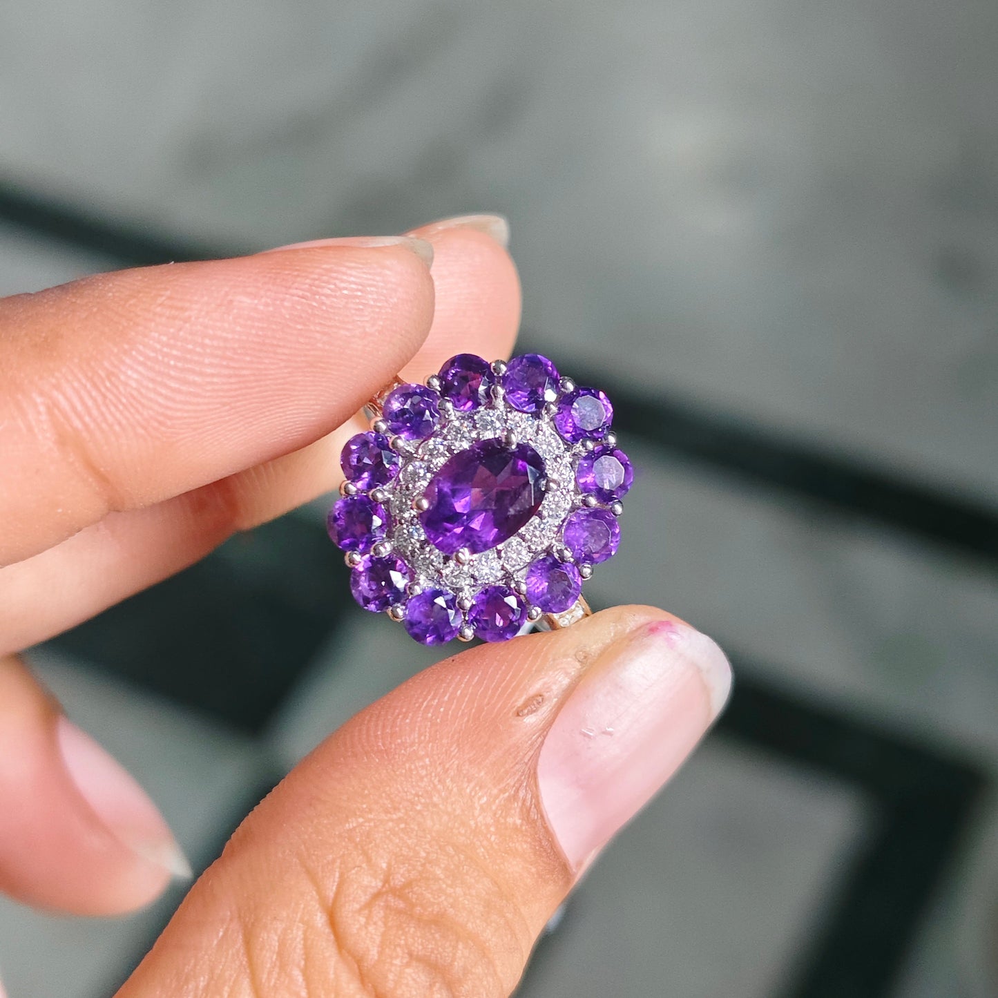 A close-up of a hand holding a cluster ring featuring a central oval purple gemstone surrounded by a halo of round purple and clear stones in a silver-toned setting, against a blurred gray background.
