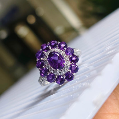 A silver ring featuring a large oval purple gemstone surrounded by a halo of round purple and clear stones, shown close-up on a white ribbed surface.