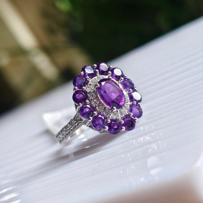 A silver ring featuring a cluster of purple amethyst gemstones, including a central oval purple stone surrounded by smaller round purple stones and a halo of clear stones, resting on a white ribbed surface.