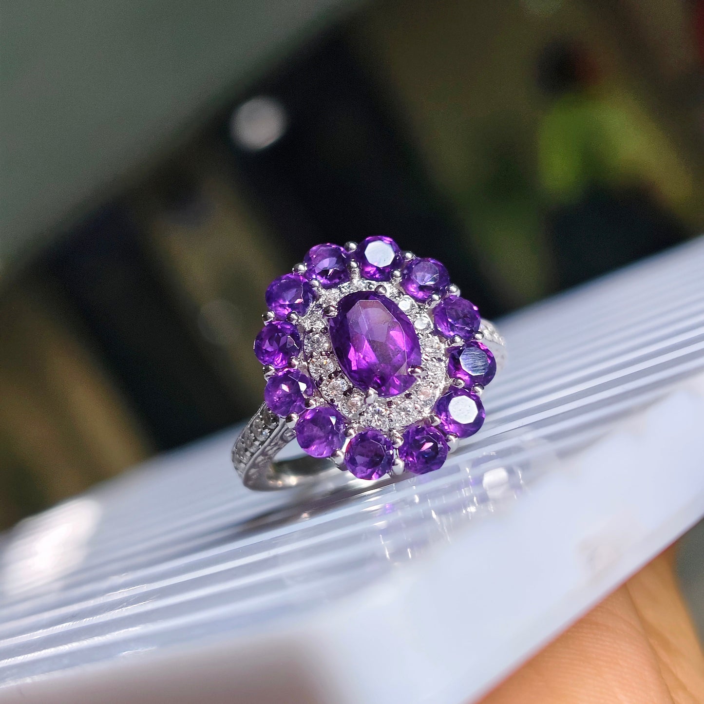 A silver ring with a central oval purple amethyst gemstone surrounded by a halo of round purple stones and small clear accent stones, shown resting on a white striped surface.