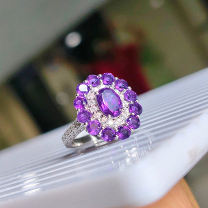 A silver ring featuring a central oval purple gemstone surrounded by a halo of round purple stones and small clear accent stones, resting on a white textured surface.