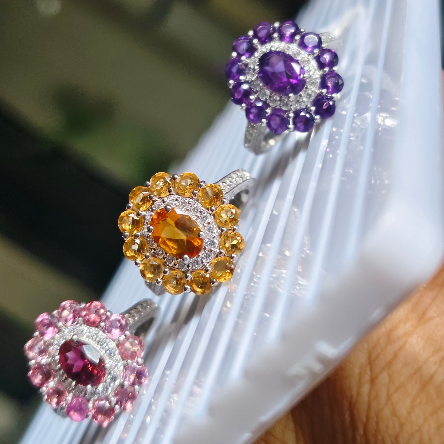 Three floral cluster rings displayed on a white ribbed surface, featuring purple, yellow-gold, and pink gemstones with silver and white metal settings.