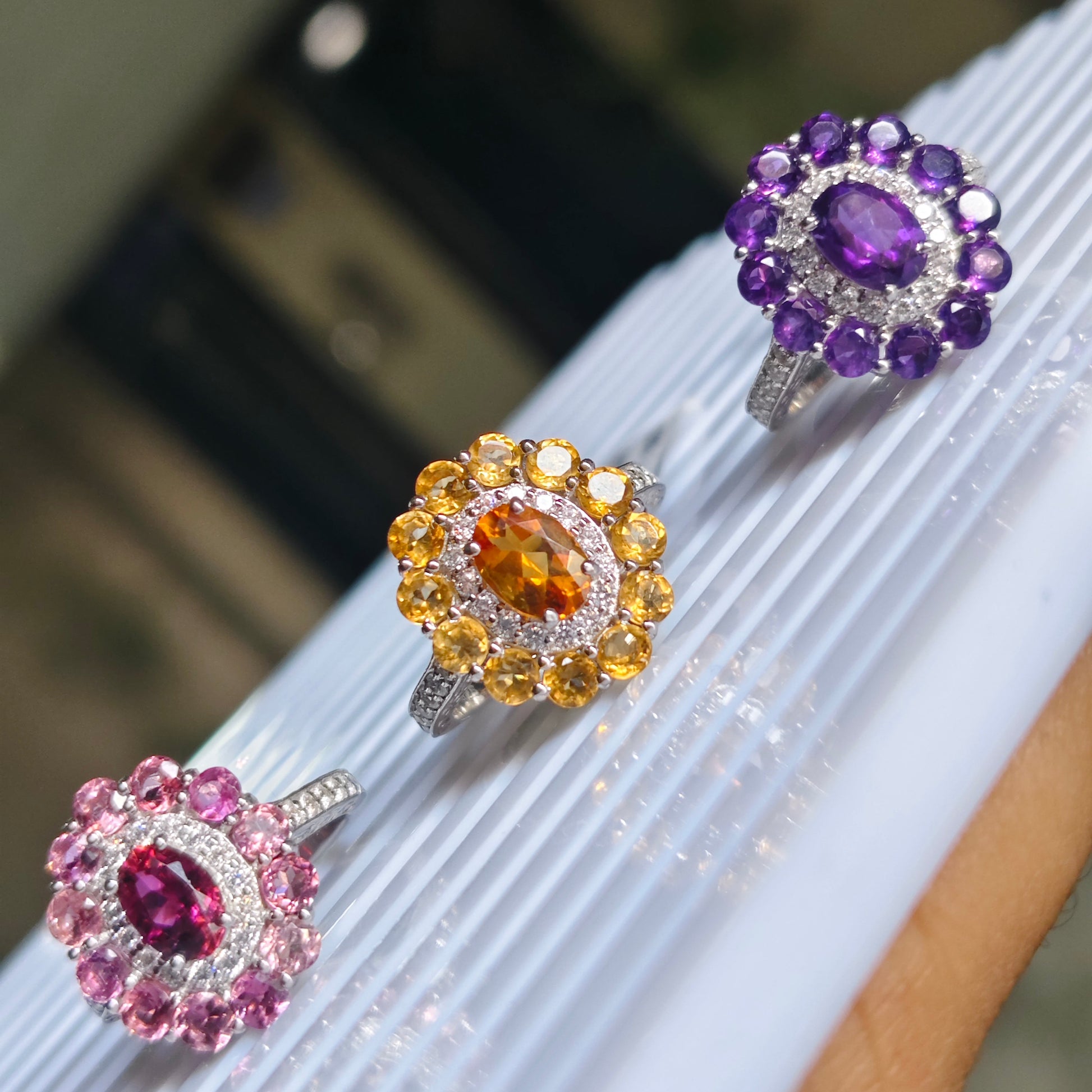 Three floral cluster rings displayed on a white ribbed surface. The rings feature purple, pink, and yellow gemstones in silver settings, with a blurred indoor background.