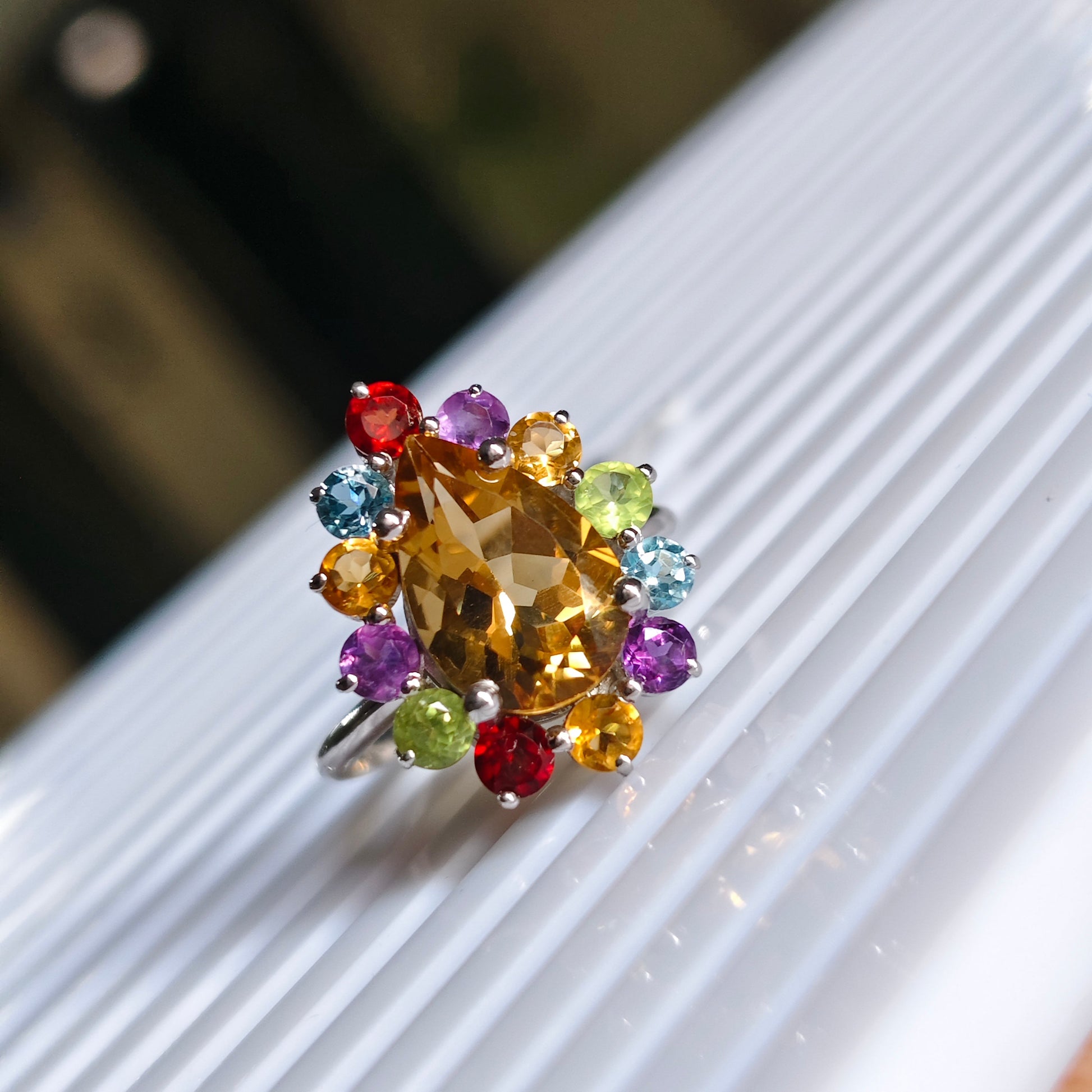 A cluster ring featuring a large central yellow-gold marquise-cut stone surrounded by a halo of smaller multicolored gemstones in red, purple, blue, green, and orange, set on a silver band and resting on a white ribbed surface.