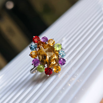 A cluster ring featuring a large central yellow-gold marquise-cut stone surrounded by a halo of smaller multicolored gemstones in red, purple, blue, green, and orange, set on a silver band and resting on a white ribbed surface.