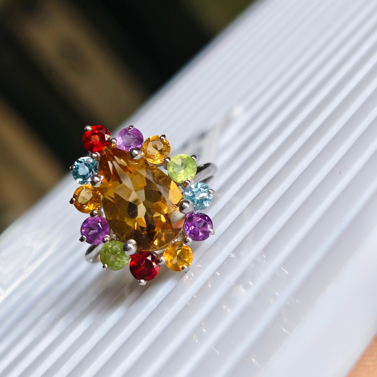 A silver ring featuring a large faceted yellow citrine center stone surrounded by a halo of small multicolored gemstones in red, purple, light blue, green, and orange, resting on a white ribbed surface.