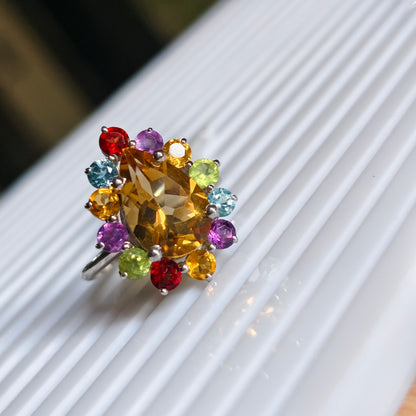 A silver ring featuring a large faceted yellow citrine center stone surrounded by a colorful halo of small multicolored gemstones, resting on a white ribbed surface.