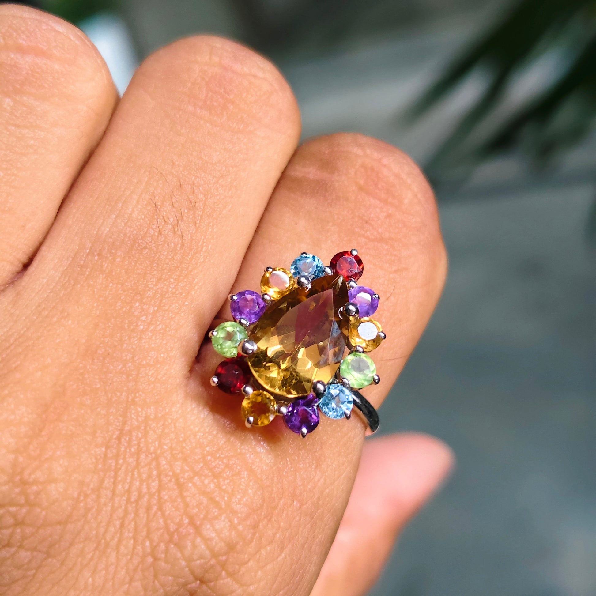 A close-up of a hand wearing a colorful gemstone ring. The ring features a central marquise-cut yellow gemstone surrounded by a cluster of small multicolored stones in purple, blue, green, orange, and red, set on a dark metal band.