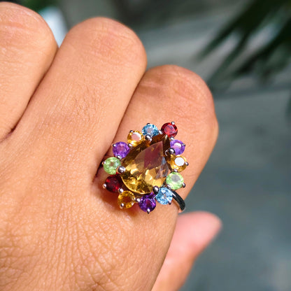 A close-up of a hand wearing a colorful gemstone ring. The ring features a central marquise-cut yellow gemstone surrounded by a cluster of small multicolored stones in purple, blue, green, orange, and red, set on a dark metal band.