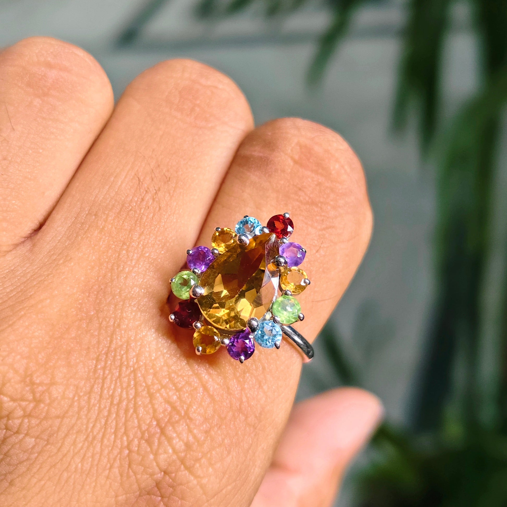 A close-up of a hand wearing a colorful gemstone cluster ring. The ring features a large yellow center stone surrounded by small multicolored stones in purple, blue, green, and red, set in a silver-toned band.