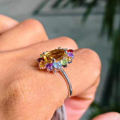 A silver ring with a large oval yellow gemstone and smaller multicolored accent stones, shown worn on a person's finger with a blurred outdoor background.