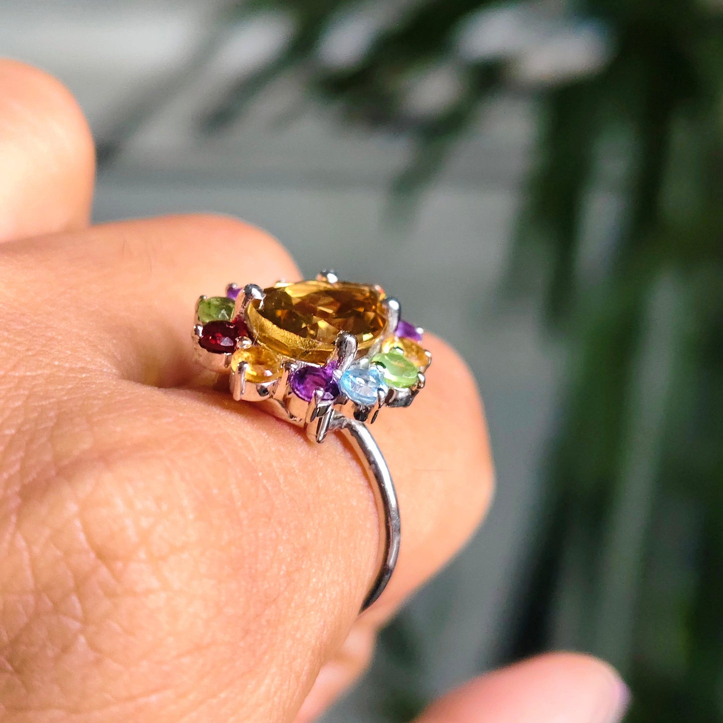 A silver ring with a large oval yellow center stone and smaller multicolored gemstones, shown worn on a person's finger against a blurred background.