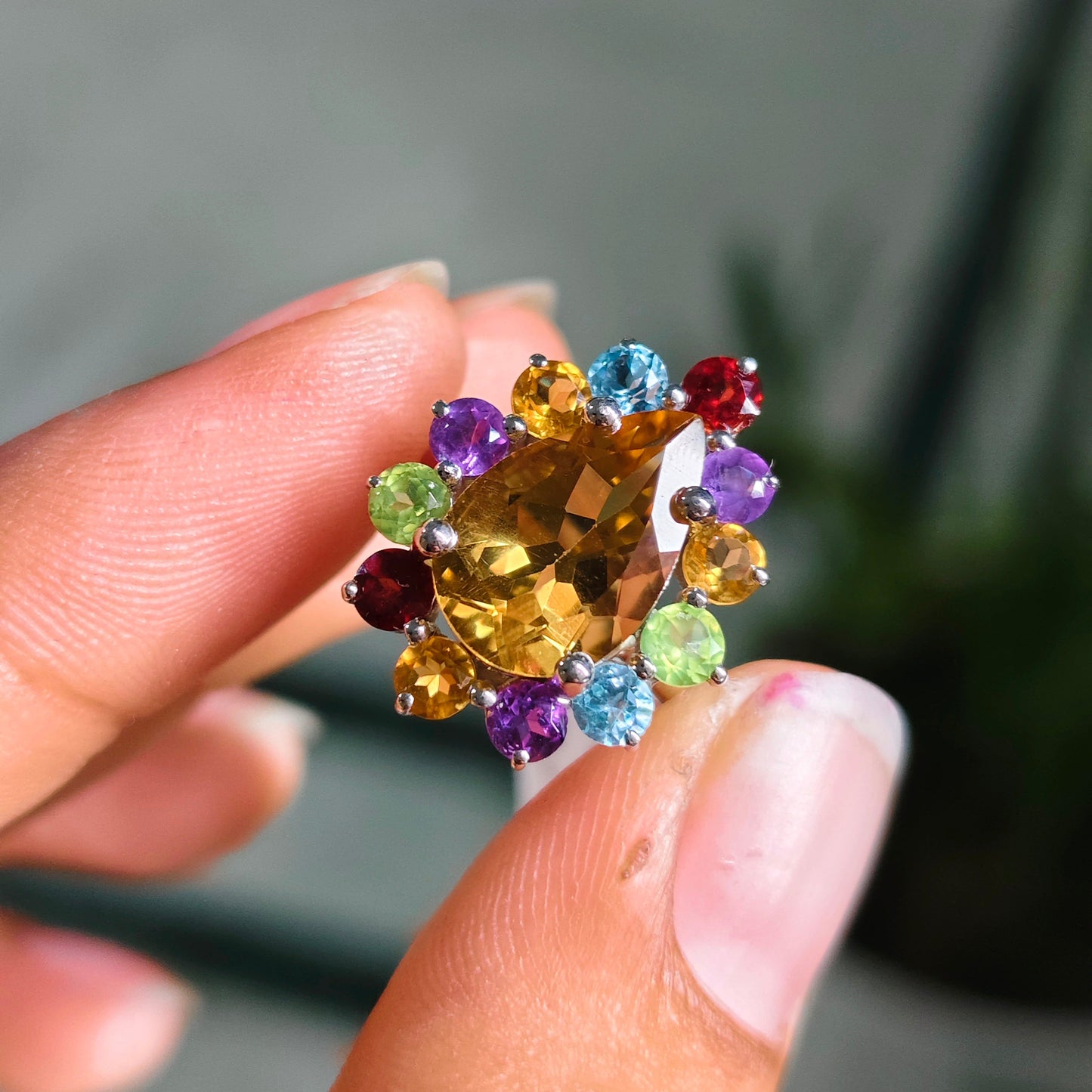 A close-up of a hand holding a cluster ring featuring a large faceted yellow citrine center stone surrounded by multiple small colorful gemstones in purple, blue, green, red, and brown.