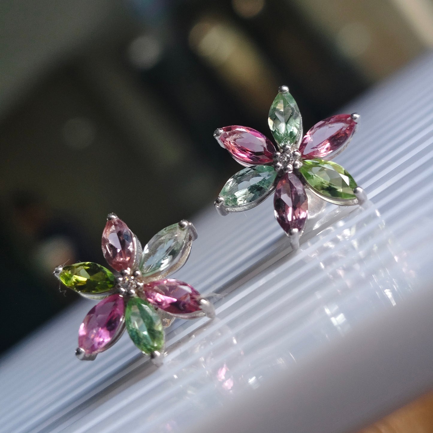 A pair of silver flower-shaped stud earrings with marquise-cut pink and green gemstones, displayed on a white ribbed surface with a blurred indoor background.