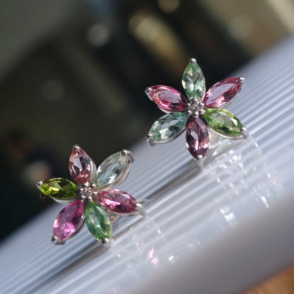 A pair of silver flower-shaped stud earrings with marquise-cut pink and green gemstones, displayed on a white ribbed surface with a blurred indoor background.