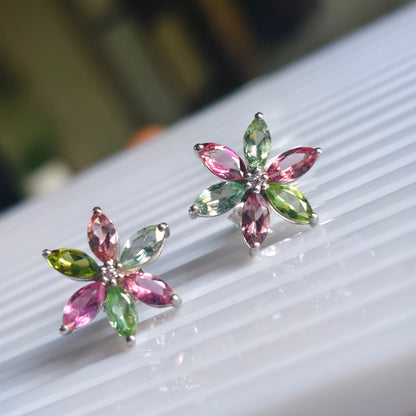 A pair of silver flower-shaped stud earrings with marquise-cut pink and green gemstones, displayed on a white ribbed surface.