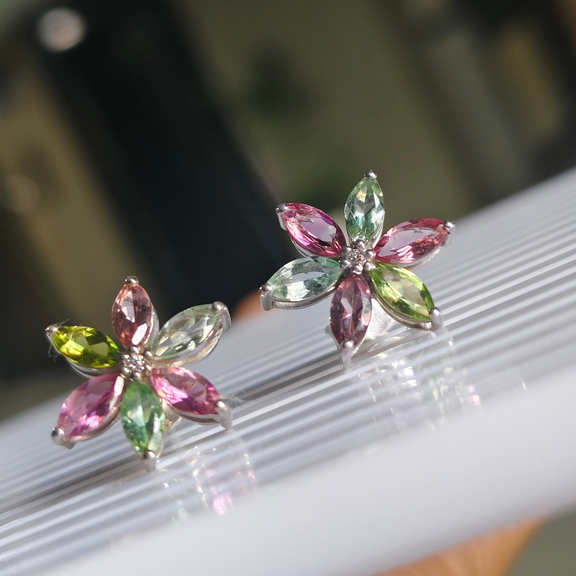 A pair of silver flower-shaped stud earrings with marquise-cut gemstones in pink, green, and yellow, resting on a white ribbed surface.