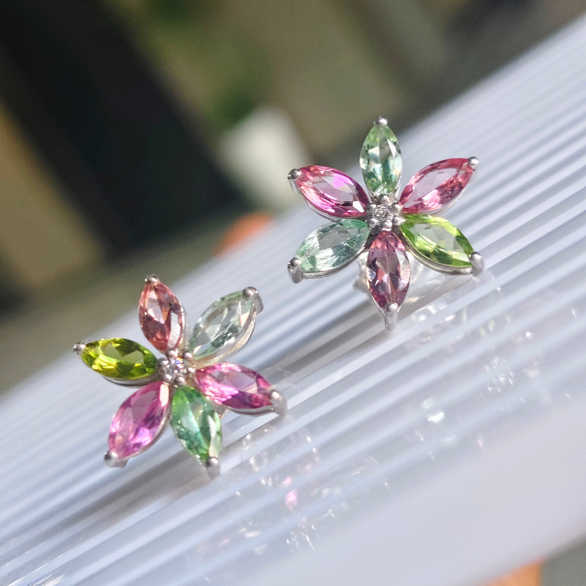 A pair of silver flower-shaped stud earrings with marquise-cut pink, green, and yellow gemstones, displayed on a white ribbed surface with a blurred indoor background.