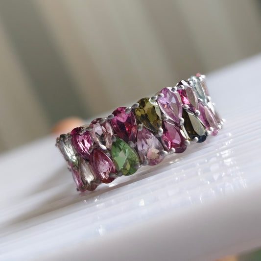 A silver band ring set with multiple marquise-cut multicolor gemstones, including pink, green, and purple, resting on a white surface.