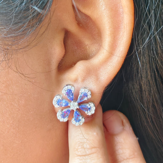 A close-up of a person's ear wearing a flower-shaped stud earring with clear rhinestones and blue iridescent centers, with a finger touching the earring.