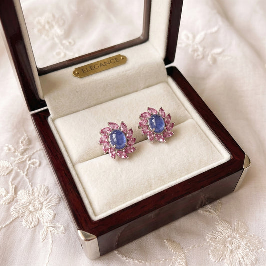 A close-up of a hand holding two silver-toned flower-shaped earrings with oval blue center stones and pink petal accents, set against a soft embroidered fabric background.