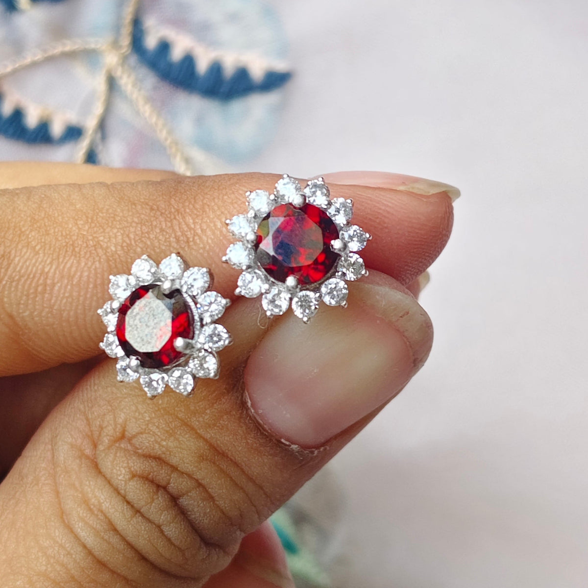 A close-up of a hand holding a pair of silver-toned stud earrings with round red center stones and small clear surrounding stones, against a soft light background with decorative floral embroidery.