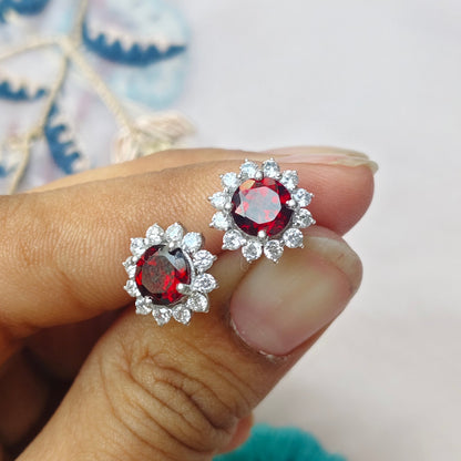 A close-up of a hand holding a pair of silver-toned stud earrings with round red gemstones surrounded by small clear stones, against a soft light background with decorative blurred elements.