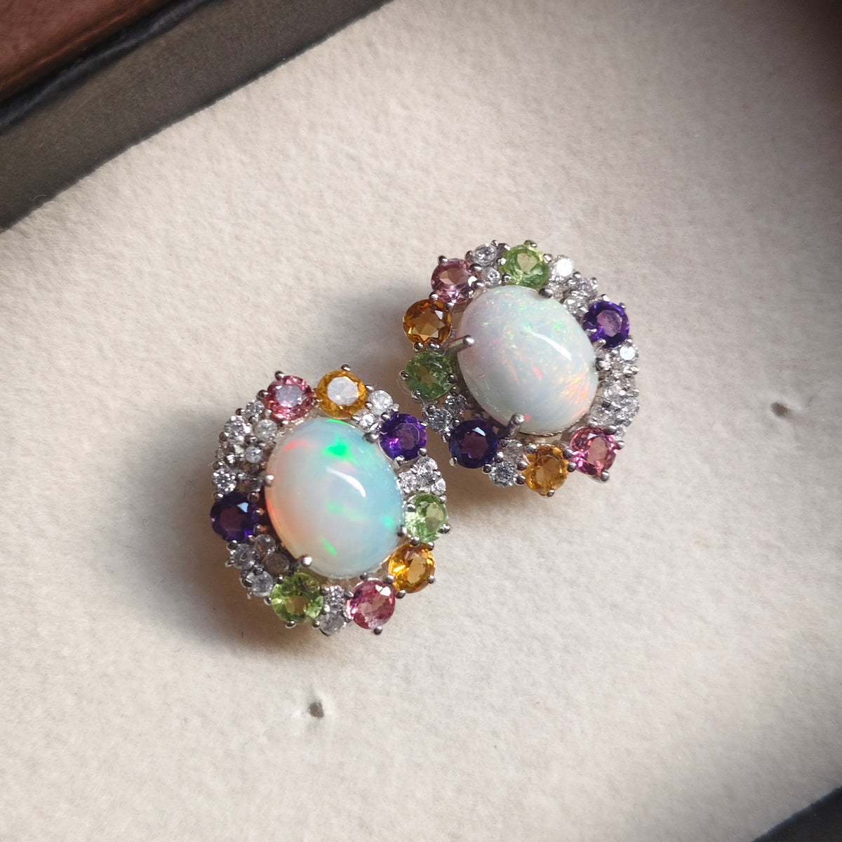 A pair of oval stud earrings with white opal centers surrounded by multicolored gemstones, displayed on a cream-colored jewelry box.